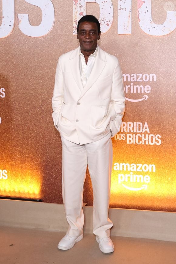 Seu Jorge aposta em um terno monocromático off-white de caimento relaxado, composto por blazer transpassado com bolsos utilitários e lapelas amplas, combinado a uma camisa acetinada levemente aberta. A calça de alfaiataria reta complementa o conjunto fluido, arrematado de forma contemporânea por tênis brancos casuais