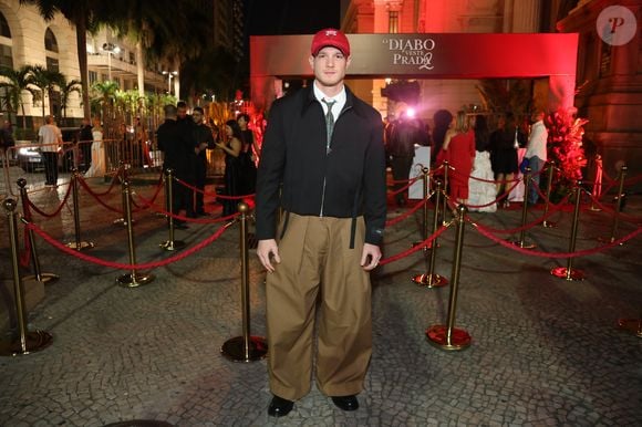 Pedro Novaes, o Léo de 'Três Graças', quebrou totalmente o protocolo formal com uma estética urbana e utilitária. Ele combinou uma jaqueta preta de corte cropped com zíper frontal e camisa branca com gravata estampada. O destaque do look foram as calças baggy de sarja cáqui, extremamente volumosas, e o boné vermelho da marca Chicago Bulls, que serviu como o ponto de cor focal da produção, finalizada com botas de sola tratorada