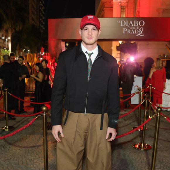 Pedro Novaes, o Léo de 'Três Graças', quebrou totalmente o protocolo formal com uma estética urbana e utilitária. Ele combinou uma jaqueta preta de corte cropped com zíper frontal e camisa branca com gravata estampada. O destaque do look foram as calças baggy de sarja cáqui, extremamente volumosas, e o boné vermelho da marca Chicago Bulls, que serviu como o ponto de cor focal da produção, finalizada com botas de sola tratorada
