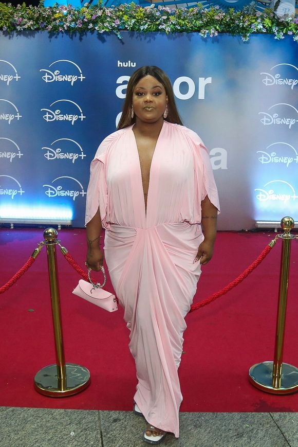 MC Carol: surge com um vestido longo em tom rosa-bebe, marcado por decote profundo em "V", mangas com fendas, drapeado frontal na cintura e bolsa de mão rosa com alça estruturada
