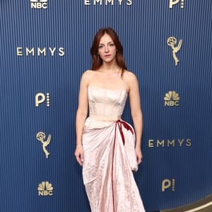 Abby Elliott ostenta um vestido rosa-claro composto por corpete estruturado estilo corset tomara que caia e saia longa drapeada. O design traz um laço de veludo bordô amarrado na cintura que adiciona contraste, enquanto a saia em tecido acetinado de efeito amassado cria textura fluida. A atriz mantém os cabelos ruivos soltos e maquiagem suave