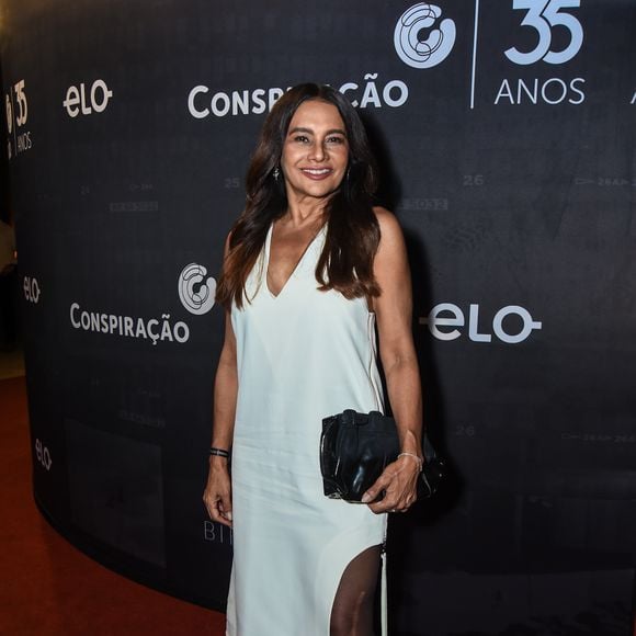 Dira Paes, a Lígia de 'Três Graças', aposta na elegância minimalista com toque sensual. A convidada surge com um vestido midi de corte reto em tom off-white, com decote em V profundo e alças largas, que valoriza a silhueta de forma sutil. A fenda lateral estratégica revela a perna com sofisticação, combinada a uma meia-calça preta fina que adiciona contraste e modernidade. Nos pés, scarpins pretos de bico fino reforçam a proposta clássica, enquanto a bolsa clutch em couro preto traz praticidade sem perder a elegância