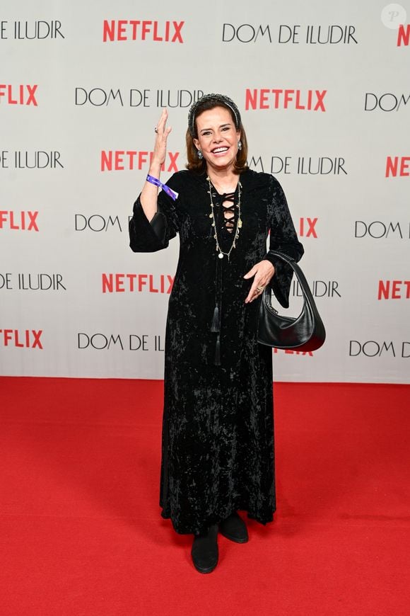 Narcisa Tamborindeguy veste um vestido longo preto texturizado com inspiração boho. A peça tem mangas 3/4 com punhos elásticos e um decote frontal com cordões entrelaçados e detalhes de metal. Ela finaliza o look com um colar longo de contas pretas e douradas, sapatos pretos fechados e segura uma bolsa de ombro preta estruturada