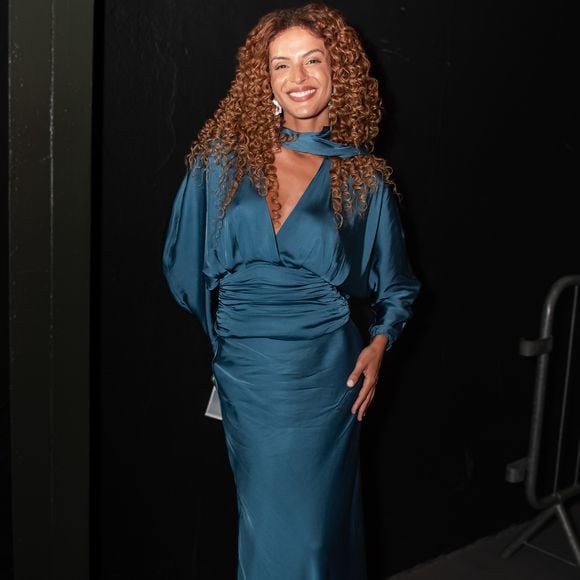 Gabriela Loran, a Viviane em 'Três Graças', brilhou em uma proposta monocromática em tom azul-petróleo, utilizando um vestido de cetim com brilho acetinado e caimento impecável. O modelo apresenta mangas longas levemente bufantes nos punhos e uma gola alta com amarração lateral, criando um drapeado sofisticado no busto. A saia fluida e a cintura marcada por uma faixa do mesmo tecido reforçam a elegância clássica da apresentadora, que optou por deixar seus cachos dourados em evidência e usar brincos geométricos prateados para iluminar o rosto