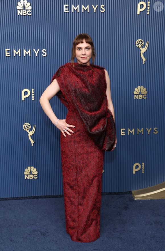 Fiona Dourif: vestido longo estruturado em tom vermelho-fechado com textura felpuda e fios mesclados em preto. A peça possui decote alto e traz um drapeado volumoso em estilo mantô no ombro esquerdo, criando uma silhueta assimétrica. A produção é complementada por franja reta, maquiagem neutra e joias prateadas discretas