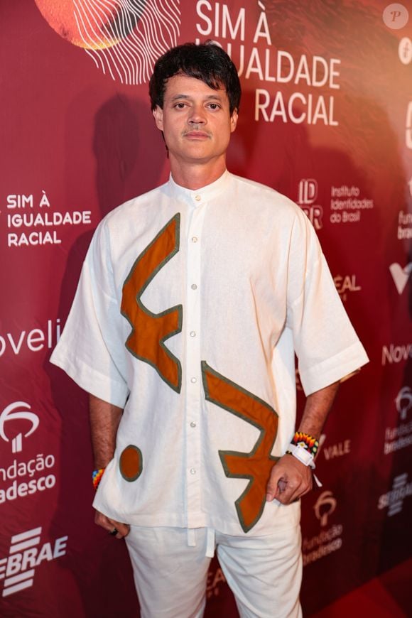 Álamo Facó escolheu um conjunto monocromático branco que se destaca pelas aplicações geométricas em tom caramelo. A camisa, de modelagem ampla com mangas 3/4 e gola padre, serve como tela para os recortes abstratos que lembram símbolos rústicos. Acompanhado de uma calça de corte reto no mesmo material, o ator adicionou um toque de cor nos pulsos com pulseiras artesanais de miçangas coloridas, mantendo uma estética leve e autoral
