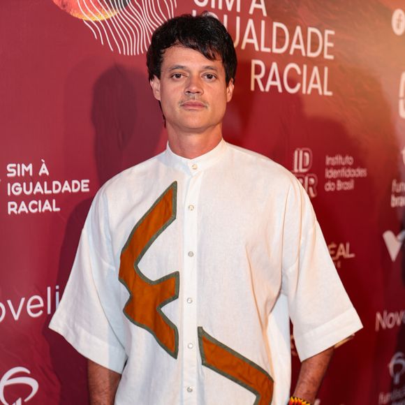 Álamo Facó escolheu um conjunto monocromático branco que se destaca pelas aplicações geométricas em tom caramelo. A camisa, de modelagem ampla com mangas 3/4 e gola padre, serve como tela para os recortes abstratos que lembram símbolos rústicos. Acompanhado de uma calça de corte reto no mesmo material, o ator adicionou um toque de cor nos pulsos com pulseiras artesanais de miçangas coloridas, mantendo uma estética leve e autoral