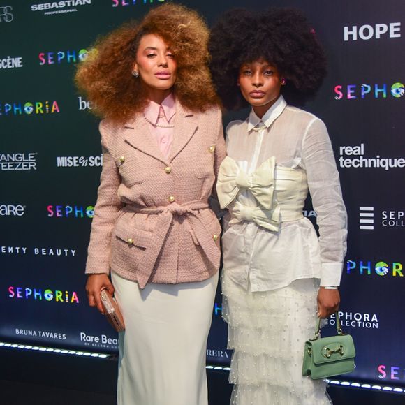 Raissa Santana e sua acompanhante trouxeram uma proposta de alfaiataria romântica e texturizada. Raissa (à esquerda) veste um blazer de tweed rosa seco com botões dourados e amarração na cintura, combinado com uma saia longa fluida em tom off-white. Já a convidada ao seu lado apresenta um visual lúdico com uma camisa branca sob um top estruturado com laços gigantes, finalizado com uma saia de camadas de tule e pérolas, acessórios em tom pastel e sapatos lilás