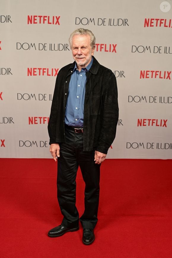 Jonas Bloch veste um blazer preto texturizado sobre uma camisa de botão azul clara de algodão. Ele completa a produção casual com calças jeans pretas de corte reto, cinto de couro preto com fivela prateada e sapatos sociais pretos de amarrar