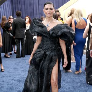 Lisa Rinna: vestido de alta-costura preto em tecido reluzente e estruturado. O modelo apresenta decote dramático com mangas volumosas e drapeados esculturais por todo o comprimento, além de uma fenda lateral profunda e cauda longa. O look é arrematado por um colar com pedra azul e scarpins pretos de bico fino
