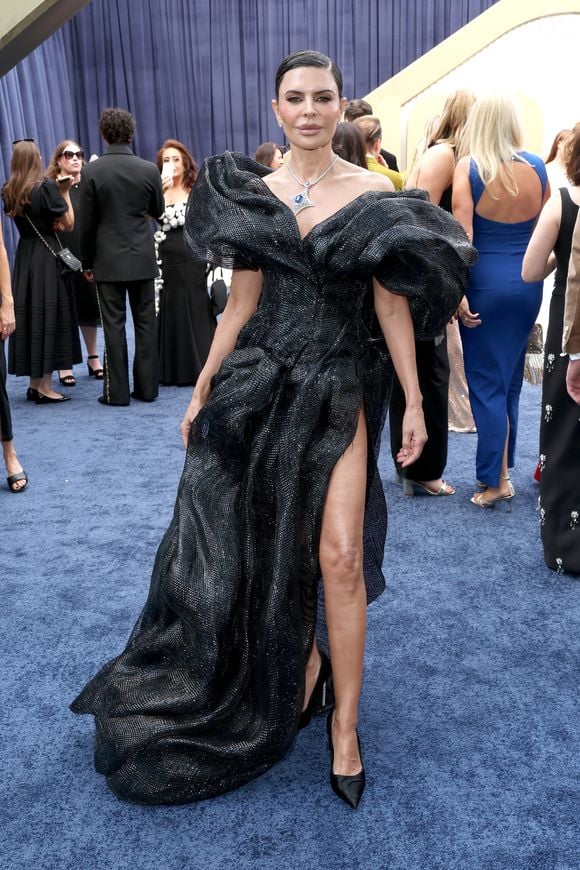 Lisa Rinna: vestido de alta-costura preto em tecido reluzente e estruturado. O modelo apresenta decote dramático com mangas volumosas e drapeados esculturais por todo o comprimento, além de uma fenda lateral profunda e cauda longa. O look é arrematado por um colar com pedra azul e scarpins pretos de bico fino