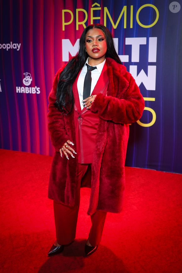 Duquesa apostou em uma estética imponente e cheia de personalidade, misturando tailoring e textura como quem diz: 'moda é poder'. O look traz um conjunto vermelho de alfaiataria impecável - blazer estruturado, camisa branca e gravata preta - que já seria impactante sozinho, mas ganha camadas de teatralidade com o longo casaco de pele fake na mesma paleta, volumoso e luxuoso