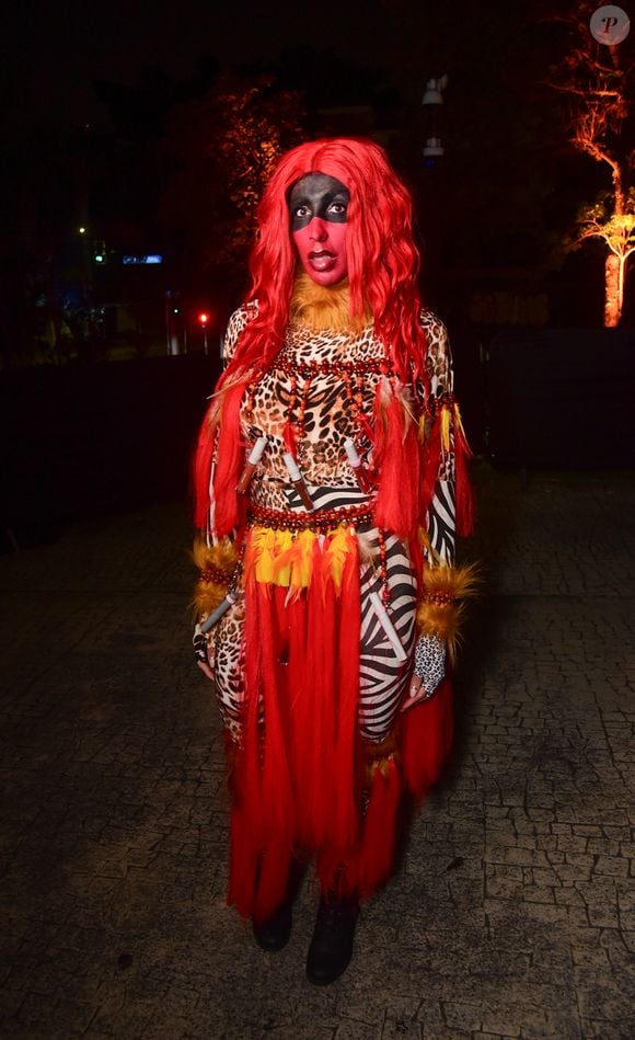 Pepita veste uma fantasia vibrante e performática em estampa animal print e tons avermelhados, inspirada na Caipora. Ela usa macacão justo com estampas mistas de onça e zebra, acompanhado por detalhes em franjas e tecidos vermelhos sobrepostos na cintura. O visual conta ainda com peruca longa em tom vermelho vivo, pintura facial temática em preto e vermelho ao redor dos olhos e detalhes acolchoados nos punhos