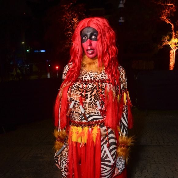 Pepita veste uma fantasia vibrante e performática em estampa animal print e tons avermelhados, inspirada na Caipora. Ela usa macacão justo com estampas mistas de onça e zebra, acompanhado por detalhes em franjas e tecidos vermelhos sobrepostos na cintura. O visual conta ainda com peruca longa em tom vermelho vivo, pintura facial temática em preto e vermelho ao redor dos olhos e detalhes acolchoados nos punhos