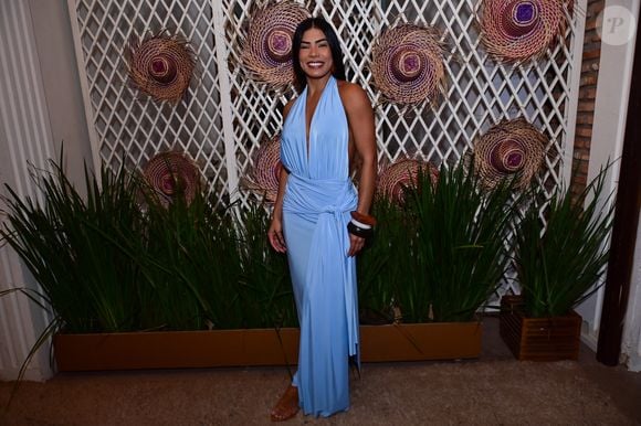 Marciele: escolheu a sofisticação fluida de um vestido longo azul-claro de decote frente única profundo e drapeado. A peça traz uma amarração em nó na linha do quadril que cria uma fenda sutil, coordenada com braceletes largos em tons de marrom e branco e sandálias de dedo rasteiras