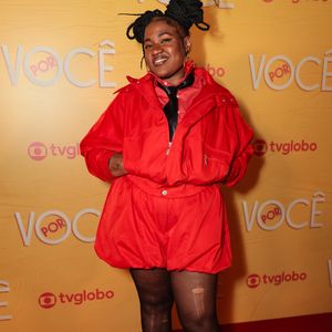 Ariane Souza exala atitude streetwear em um conjunto monocromático vermelho composto por jaqueta estilizada de gola alta e bermuda de modelagem balão com cós elástico. O figurino traz gravata preta fina por baixo da gola, meia-calça preta semitransparente com pequenos rasgos e botas de cano alto