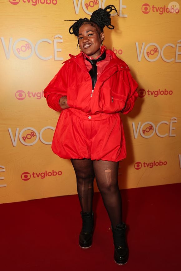 Ariane Souza exala atitude streetwear em um conjunto monocromático vermelho composto por jaqueta estilizada de gola alta e bermuda de modelagem balão com cós elástico. O figurino traz gravata preta fina por baixo da gola, meia-calça preta semitransparente com pequenos rasgos e botas de cano alto