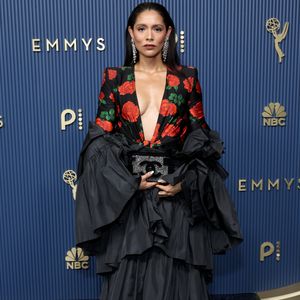 Miranda Rae Mayo surge em uma produção dramática de saia preta em camadas de babados plissados e top de mangas longas com ombros estruturados e estampa floral de rosas vermelhas. O decote profundo em 'V' contrasta com o volume exagerado da peça e da clutch preta com logo brilhante. Os cabelos aparecem alinhados para trás em efeito wet