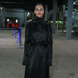 Alanis Guillen: usa um trench coat longo preto acetinado de gola alta ajustado na cintura por amarração, combinado com bolsa de ombro e botas pretas de estilo tabi