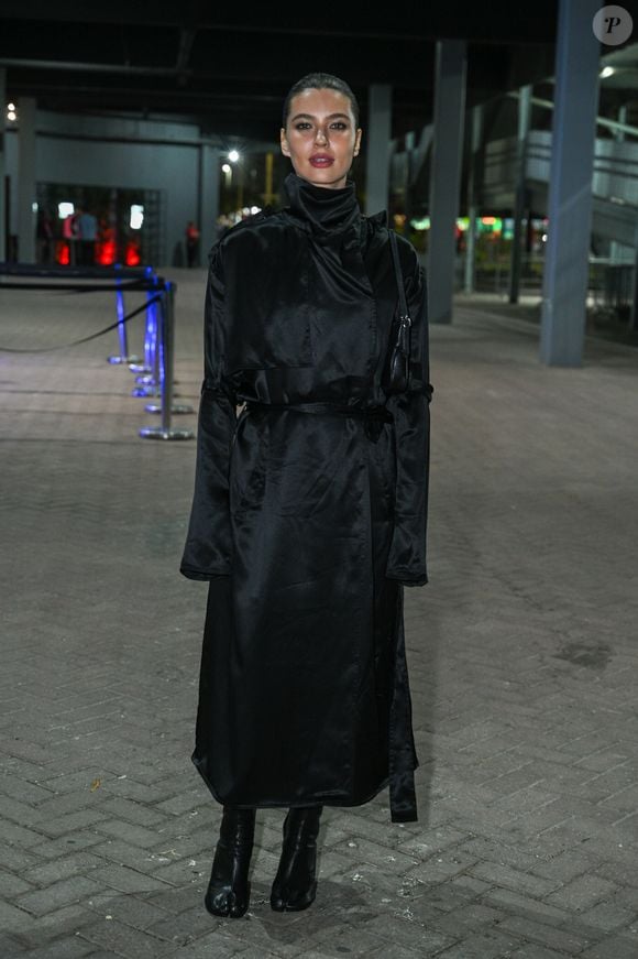Alanis Guillen: usa um trench coat longo preto acetinado de gola alta ajustado na cintura por amarração, combinado com bolsa de ombro e botas pretas de estilo tabi