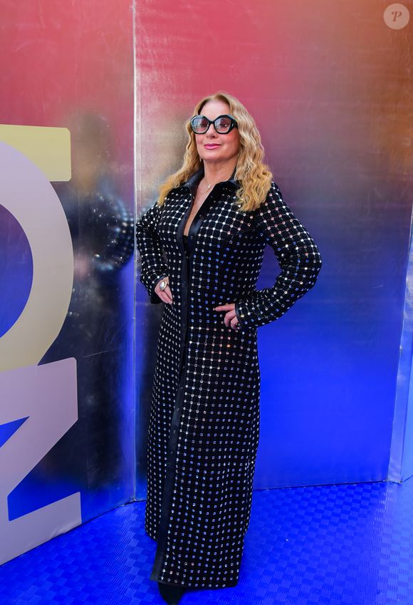 Vera Fischer: esbanjando elegância e modernidade, Vera Fischer utilizou um sobretudo longo de couro preto que funciona como a peça central do look. O casaco apresenta uma padronagem quadriculada formada por pequenas aplicações metálicas ou cristais refletivos em toda a sua extensão, criando um efeito de brilho pontuado. Por baixo, nota-se uma base preta lisa