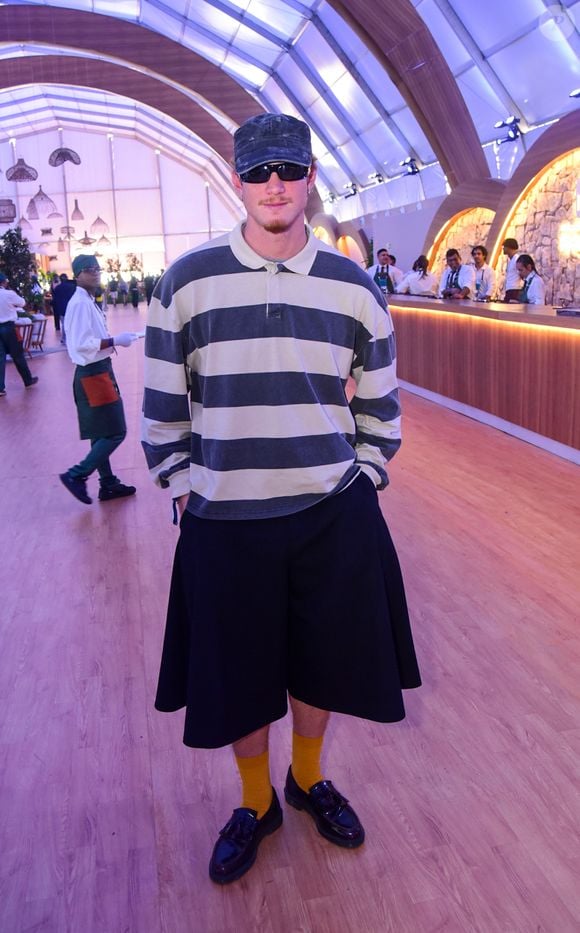 Pedro Novaes aposta em uma camisa polo de mangas compridas e modelagem oversized com listras horizontais largas em cinza e off-white, combinada com uma bermuda de alfaiataria preta super ampla de caimento plissado. O toque moderno fica por conta do boné estonado, óculos escuros com lente espelhada, meias altas na cor mostarda e mocassins pretos envernizados