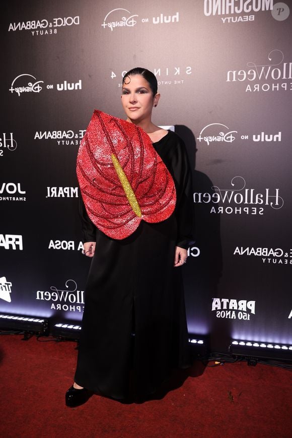 Ju Ferraz: o look de alfaiataria preta traz um vestido longo com mangas compridas e fenda central frontal, combinada com sapatos de plataforma preta. O elemento principal da peça é uma grande aplicação frontal em formato de antúrio vermelho, totalmente bordada com brilhos e lantejoulas, com o espádice amarelo em destaque. A beleza traz cabelo preso bem alinhado com uma mecha modelada na testa e maquiagem com foco nos olho