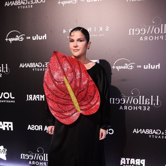 Ju Ferraz: o look de alfaiataria preta traz um vestido longo com mangas compridas e fenda central frontal, combinada com sapatos de plataforma preta. O elemento principal da peça é uma grande aplicação frontal em formato de antúrio vermelho, totalmente bordada com brilhos e lantejoulas, com o espádice amarelo em destaque. A beleza traz cabelo preso bem alinhado com uma mecha modelada na testa e maquiagem com foco nos olho