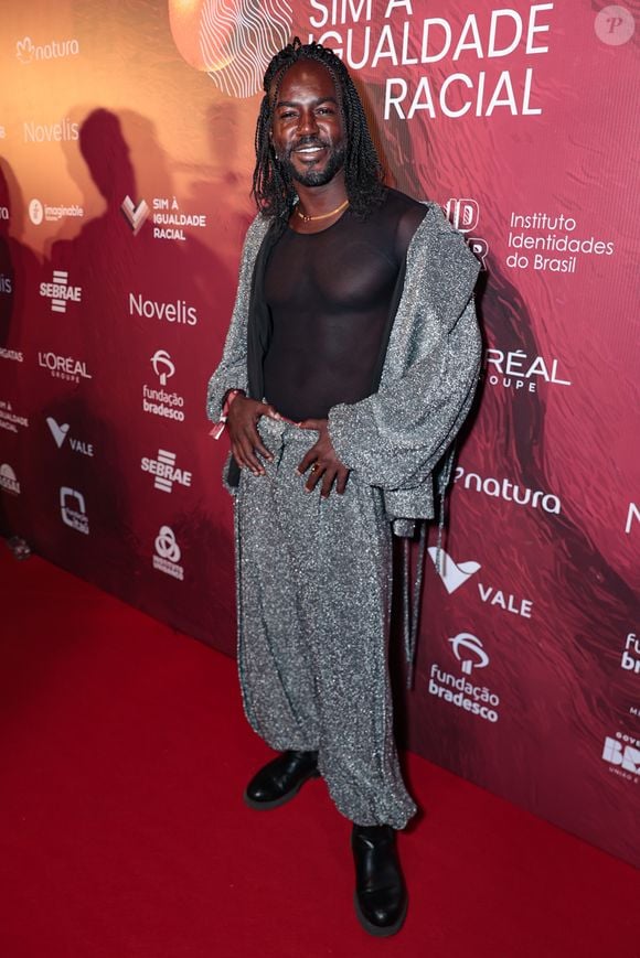 Jonathan Azevedo apostou no brilho e na fluidez para o tapete vermelho. O ator veste um conjunto de calça oversized e casaco tipo jaqueta, ambos confeccionados em um tecido prateado com textura de paetês miúdos que garantem movimento. Por baixo, uma blusa de tule preto com transparência revela a silhueta, trazendo um toque de sensualidade