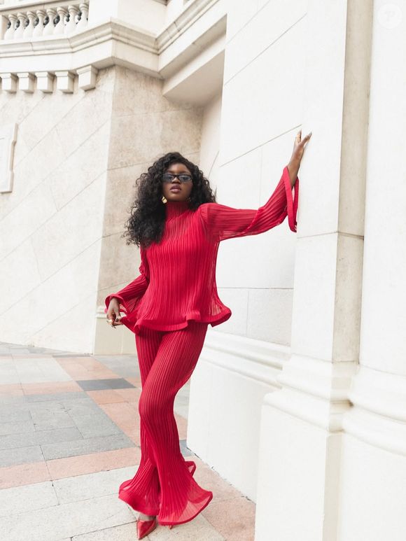 Kenya Sade apareceu como um cometa vermelho! Seu conjunto plissado all red combina blusa de mangas longas com ondas dramáticas e calça boca de sino escultural, que cria movimento com o vento. Os sapatos vermelhos, óculos escuros modernos, brincos dourados e cabelo volumoso transformam o visual em um manifesto de confiança. É aquele tipo de look que diz tudo antes mesmo de a artista abrir a boca