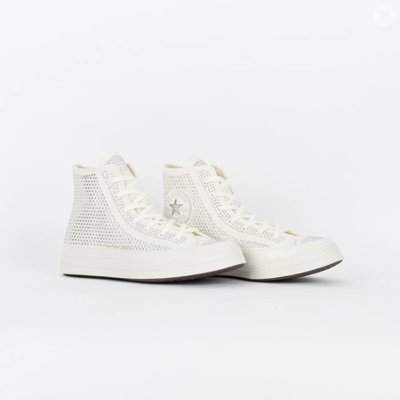 O Chuck 70 Hi aparece na cor branca, com cano alto, bico arredondado, fechamento frontal por amarração, palmilha com logo e aplicações de cristais Swarovski, criando uma versão glamourosa do clássico