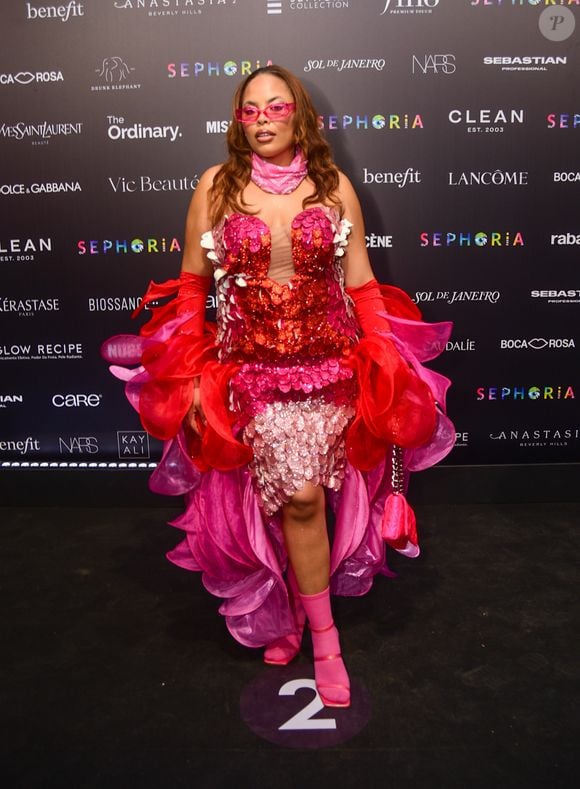 Rita Carreira optou por um visual maximalista e floral, vestindo uma peça estruturada que simulava camadas de pétalas em tons de vermelho, rosa e lilás, decorada com paetês. A modelo elevou a complexidade do look com luvas vermelhas de babados volumosos, uma bandana rosa no pescoço e óculos de sol translúcidos, criando uma composição rica em camadas e texturas