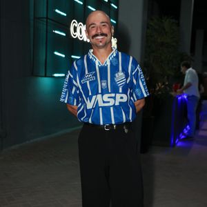 Ed Gama surgiu com uma proposta esportiva retrô cheia de presença ao vestir uma camisa vintage azul e branca do CSA (Centro Sportivo Alagoano) colocada por dentro da calça. A composição seguiu elegante com uma calça social preta de modelagem ampla, cinto preto e sapatos de sola tratorada. O visual misturou a cultura do futebol com a alfaiataria urbana contemporânea