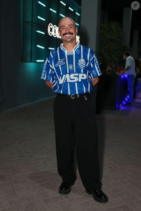 Ed Gama surgiu com uma proposta esportiva retrô cheia de presença ao vestir uma camisa vintage azul e branca do CSA (Centro Sportivo Alagoano) colocada por dentro da calça. A composição seguiu elegante com uma calça social preta de modelagem ampla, cinto preto e sapatos de sola tratorada. O visual misturou a cultura do futebol com a alfaiataria urbana contemporânea