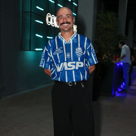 Ed Gama surgiu com uma proposta esportiva retrô cheia de presença ao vestir uma camisa vintage azul e branca do CSA (Centro Sportivo Alagoano) colocada por dentro da calça. A composição seguiu elegante com uma calça social preta de modelagem ampla, cinto preto e sapatos de sola tratorada. O visual misturou a cultura do futebol com a alfaiataria urbana contemporânea