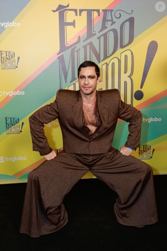 O visual de Rainer é todo construído em cima do exagero proposital. Ele escolheu um terno de tecido marrom texturizado, com blazer estruturado e calça de modelagem extra larga, quase dramática, que alonga a silhueta e dá movimento à produção. Debaixo do paletó, nada de camisa careta: ele apareceu com o peito à mostra e uma camisa aberta no mesmo tom, reforçando o ar sensual e provocador do look