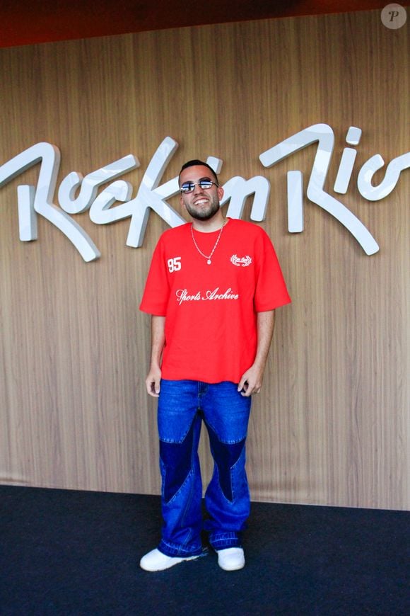 Lucas Rangel investe em uma camiseta oversized vermelha de tom vibrante com estética esportiva e estampas em estilo college. Ele combina a peça com calça jeans de lavagem média apresentando recortes geométricos em jeans escuro nos joelhos, arrematando a produção com tênis branco casual e correntaria discreta no pescoço