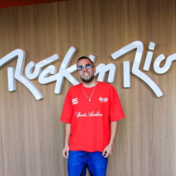 Lucas Rangel investe em uma camiseta oversized vermelha de tom vibrante com estética esportiva e estampas em estilo college. Ele combina a peça com calça jeans de lavagem média apresentando recortes geométricos em jeans escuro nos joelhos, arrematando a produção com tênis branco casual e correntaria discreta no pescoço