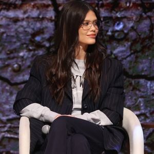 Por baixo, a atriz Vitória Strada quebra a seriedade da alfaiataria com uma camiseta de moletom cinza mescla com estampa de estilo universitário ("Hollywood"). O detalhe do cordão branco aparente na cintura reforça essa pegada esportiva