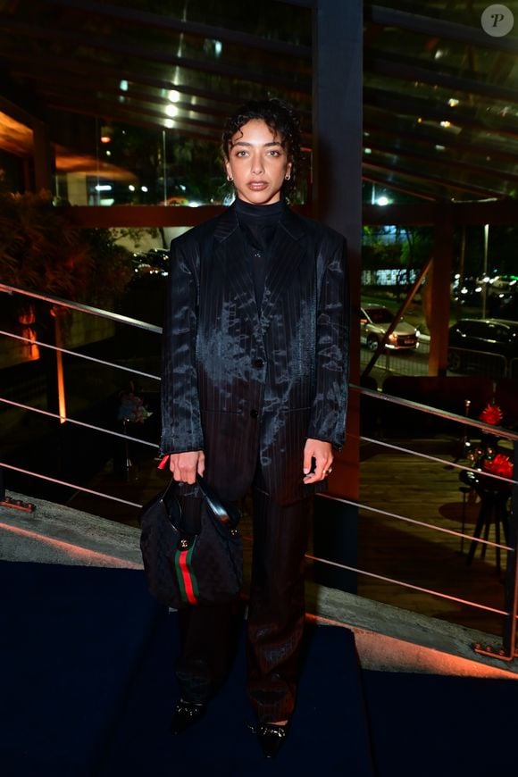 Alice Carvalho: apostou em um terno de alfaiataria oversized em tom escuro (preto/cinza) com estampa risca-de-giz e efeito manchado/texturizado em tom mais claro. O conjunto é composto por blazer amplo abotoado e calça reta, combinado com blusa de gola alta preta por baixo. Nos acessórios, carrega uma bolsa de ombro com a icônica faixa listrada verde e vermelha com monogramas e usa sapatos pretos com fivelas prateadas