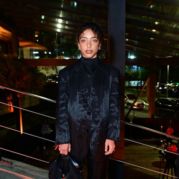 Alice Carvalho: apostou em um terno de alfaiataria oversized em tom escuro (preto/cinza) com estampa risca-de-giz e efeito manchado/texturizado em tom mais claro. O conjunto é composto por blazer amplo abotoado e calça reta, combinado com blusa de gola alta preta por baixo. Nos acessórios, carrega uma bolsa de ombro com a icônica faixa listrada verde e vermelha com monogramas e usa sapatos pretos com fivelas prateadas