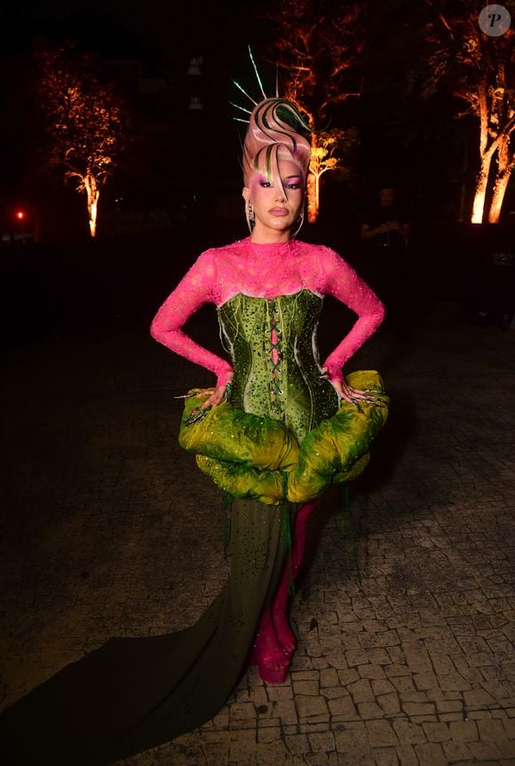 Priscila Senna veste uma criação escultural e colorida em tons de verde e rosa. O look é composto por uma blusa justa de renda rosa choque combinada a um corset verde estruturado com amarração frontal, adornado por um elemento acolchoado volumoso em formato de folha ao redor do quadril. O visual se completa com uma saia verde longa de caimento assimétrico com cauda, meia e sapatos na cor rosa e um penteado futurista estilizado