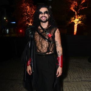 Vitor Di Castro em uma produção dramática e rock-chic. O visual conta com uma regata preta transparente de tecido telado com padrões, cruzada por tiras de couro preto que seguram uma estrela vermelha no peito e um adereço de ombro volumoso de penas pretas. Ele usa calças pretas de corte reto, botas pretas de sola robusta e braceletes de couro vermelho nos punhos. A maquiagem é pesada com olhos bem pretos e cabelo longo cacheado solto