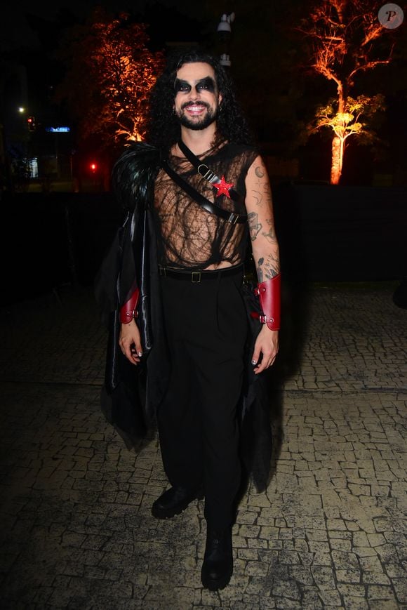 Vitor Di Castro em uma produção dramática e rock-chic. O visual conta com uma regata preta transparente de tecido telado com padrões, cruzada por tiras de couro preto que seguram uma estrela vermelha no peito e um adereço de ombro volumoso de penas pretas. Ele usa calças pretas de corte reto, botas pretas de sola robusta e braceletes de couro vermelho nos punhos. A maquiagem é pesada com olhos bem pretos e cabelo longo cacheado solto