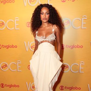 Livia Silva exibe um visual conceitual monocromático tom de off-white, composto por top estilo sutiã de renda com detalhes estruturados e aplicação de plumas no decote. A peça é combinada com saia longa de cintura alta com sobreposição geométrica e bolsos volumosos nas laterais