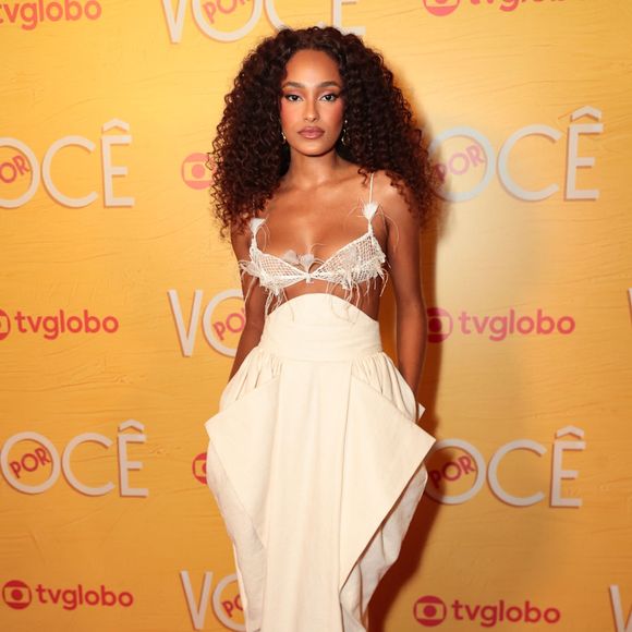 Livia Silva exibe um visual conceitual monocromático tom de off-white, composto por top estilo sutiã de renda com detalhes estruturados e aplicação de plumas no decote. A peça é combinada com saia longa de cintura alta com sobreposição geométrica e bolsos volumosos nas laterais