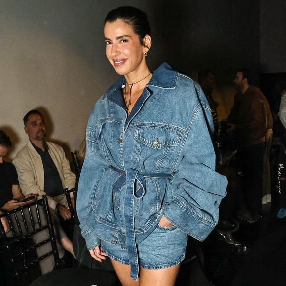 Camila Coutinho traz uma proposta total denim moderna, composta por uma jaqueta jeans oversized de lavagem azul-média com mangas volumosas e amarração na cintura pelo próprio tecido, sobreposta a um mini short jeans correspondente. Para criar contraste, o look foi finalizado com scarpins brancos de salto alto e tira no tornozelo