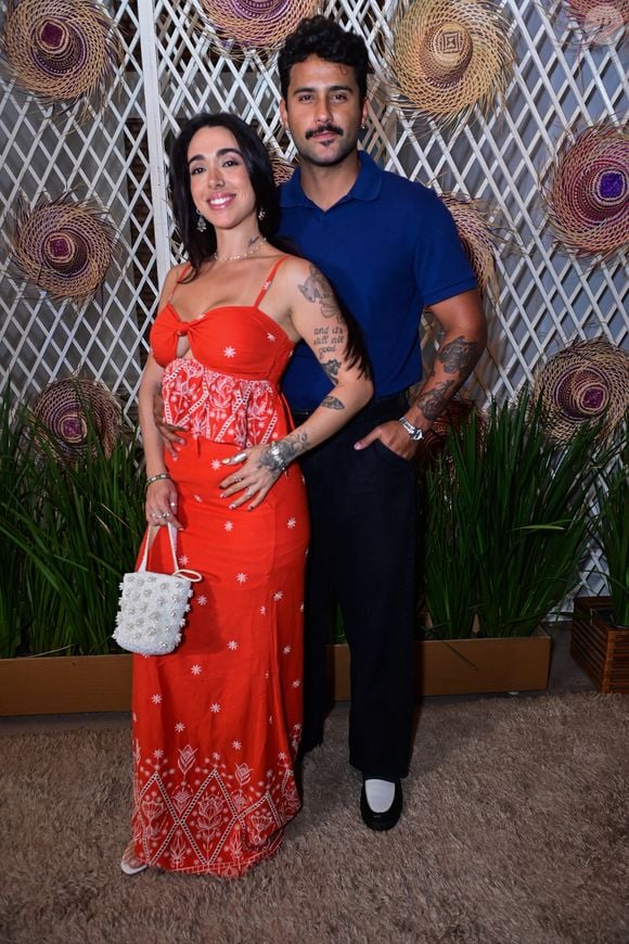 Lucas Pizane e Giovanna Lima: Giovanna elegeu um vestido longo vermelho com estampas florais brancas, recorte frontal cut-out e amarração no busto, combinado a uma bolsa de mão texturizada em pérolas. Lucas Pizane harmonizou com uma camisa polo azul-marinho lisa, calça preta de corte reto e sapatos mocassim bicolores em preto e branco