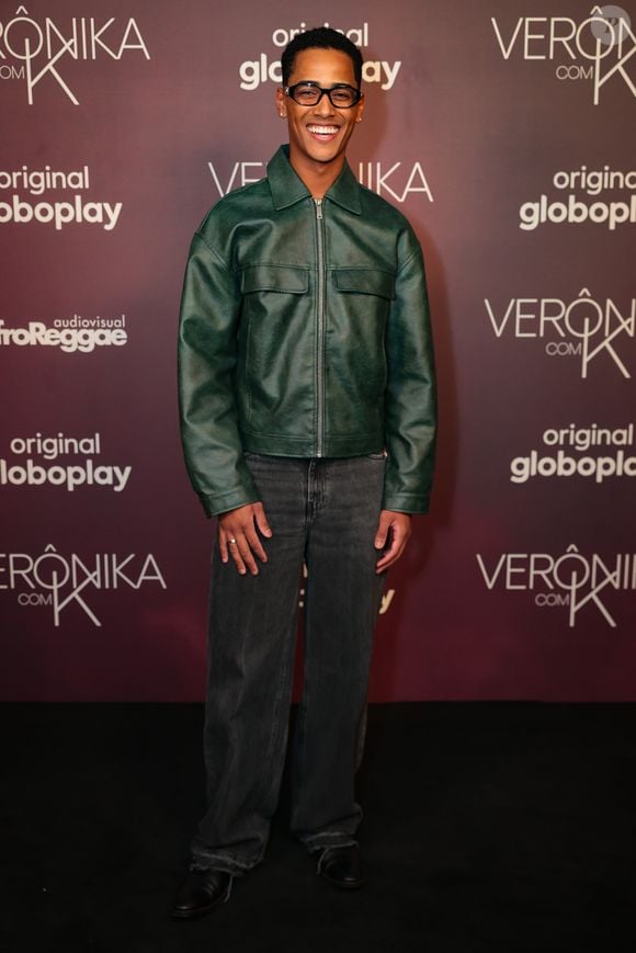 Ronald Sotto: o ator escolheu uma jaqueta de couro verde-militar com fechamento em zíper frontal e gola dobrada tradicional. A peça superior é combinada com calça jeans de lavagem cinza-escura/grafite de modelagem reta e barra desfiada. O visual moderno é complementado por óculos de armação preta marcante e sapatos sociais pretos