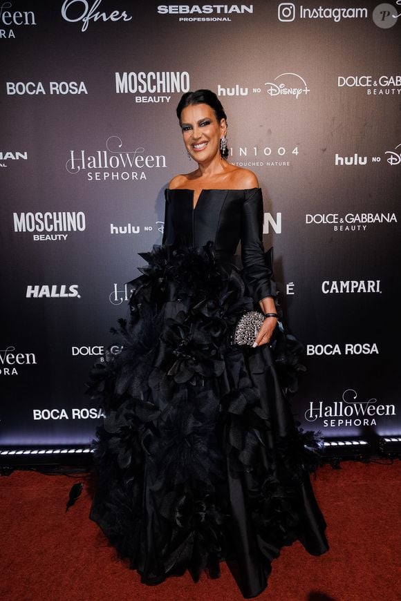 Andréia Guimarães veste um vestido de gala preto dramático com decote ombro a ombro estruturado e fenda central discreta no busto. A peça se destaca pela saia volumosa e rodada rica em aplicações tridimensionais de grandes flores e texturas em relevo por toda a extensão, combinada com clutch prateada rendada, brincos pendentes e cabelos presos num coque polido